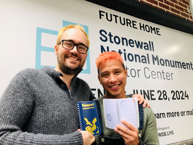 Smiling visitors pose with their NPS Passport program books in front of the brick facade of the future Stonewall National Monument Visitor Center