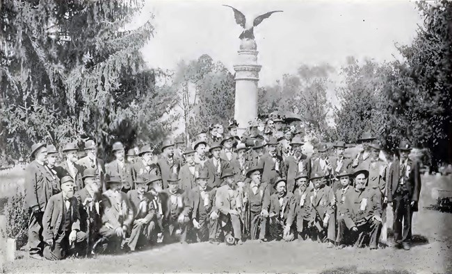 A group of older men in suits stand or kneel infront of a monument topped by an eagle.