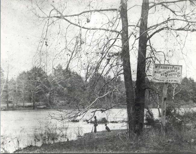 A man stands on a riverbank next a sign marking McFadden's Ford.