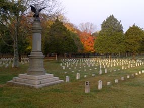 A pillar shaped monument with a bronze eagle on top stands next to rows of national cemetery headstones.