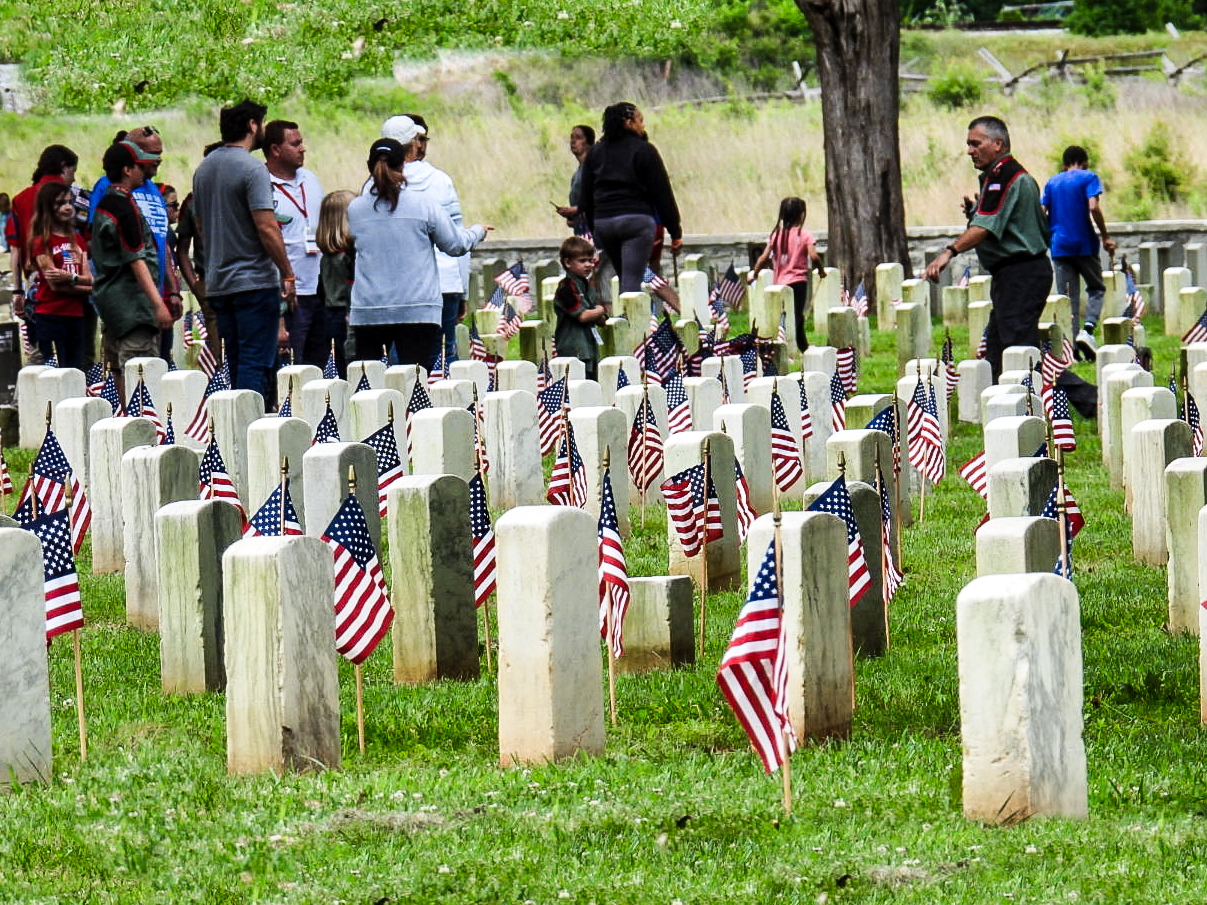 Adults and children place American flags in front of headstones aligned in rows.