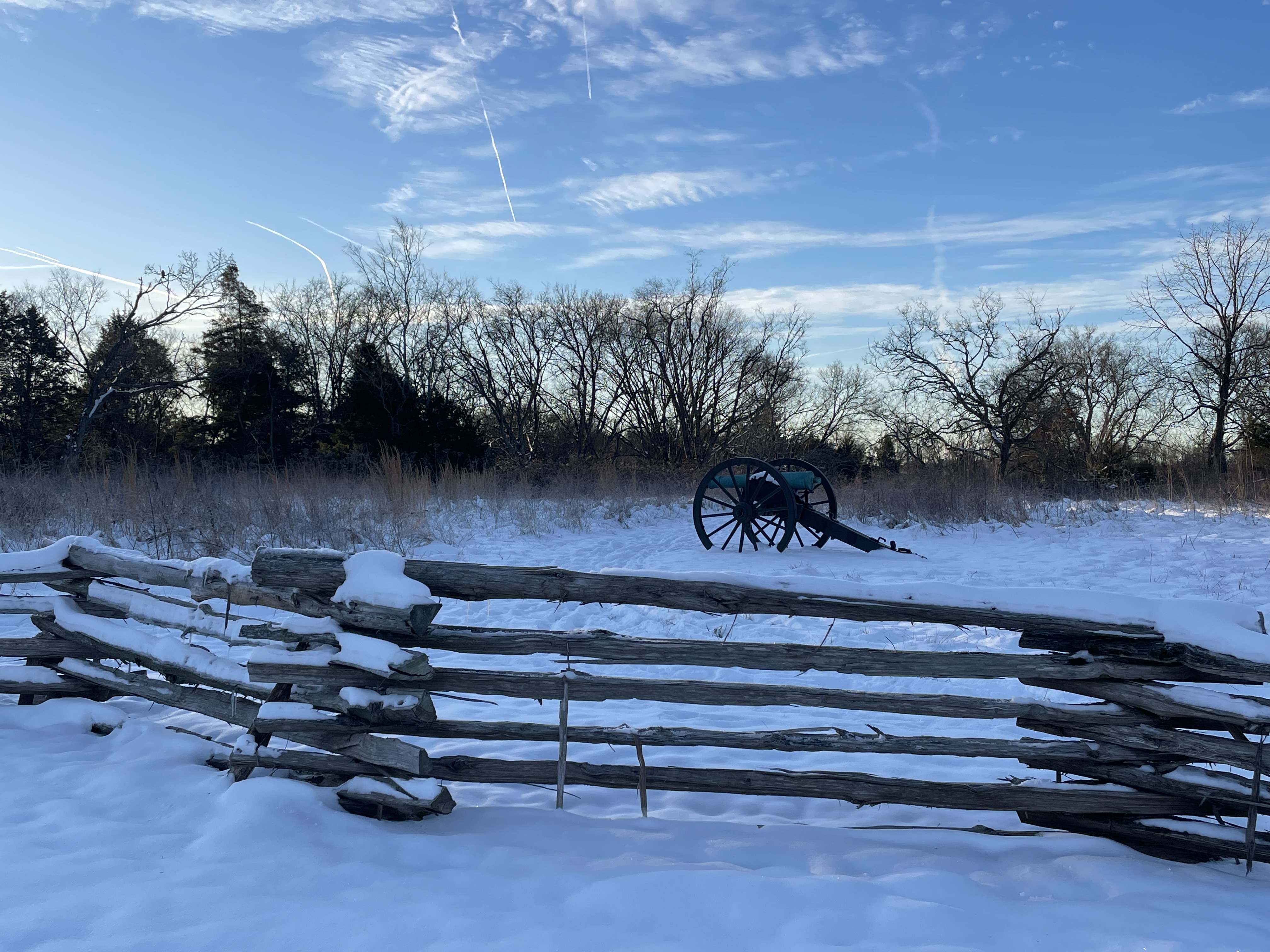 Snow covers a field bordered by a rail fence. A cannon sits in the field.