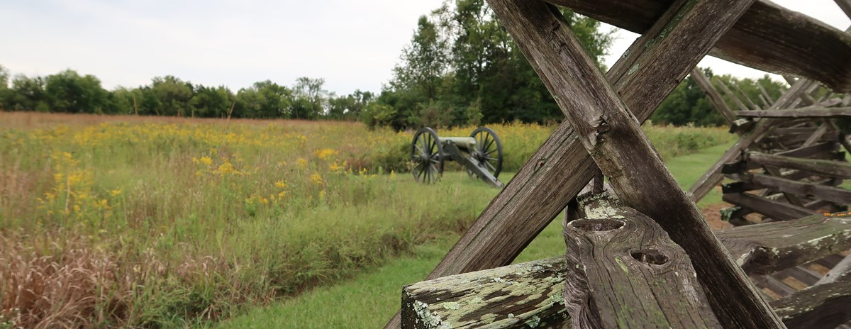 A Civil War cannon sits in a brown grass field behind a snake rail fence.