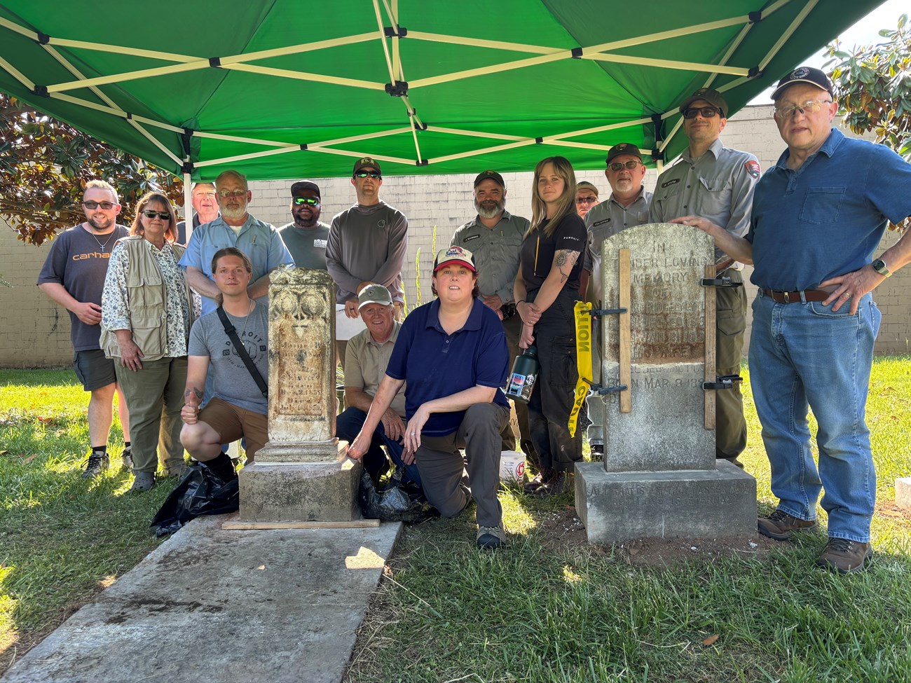 Group of park staff and volunteers standing under a green canopy around two historic gravestones during a cemetery preservation project.