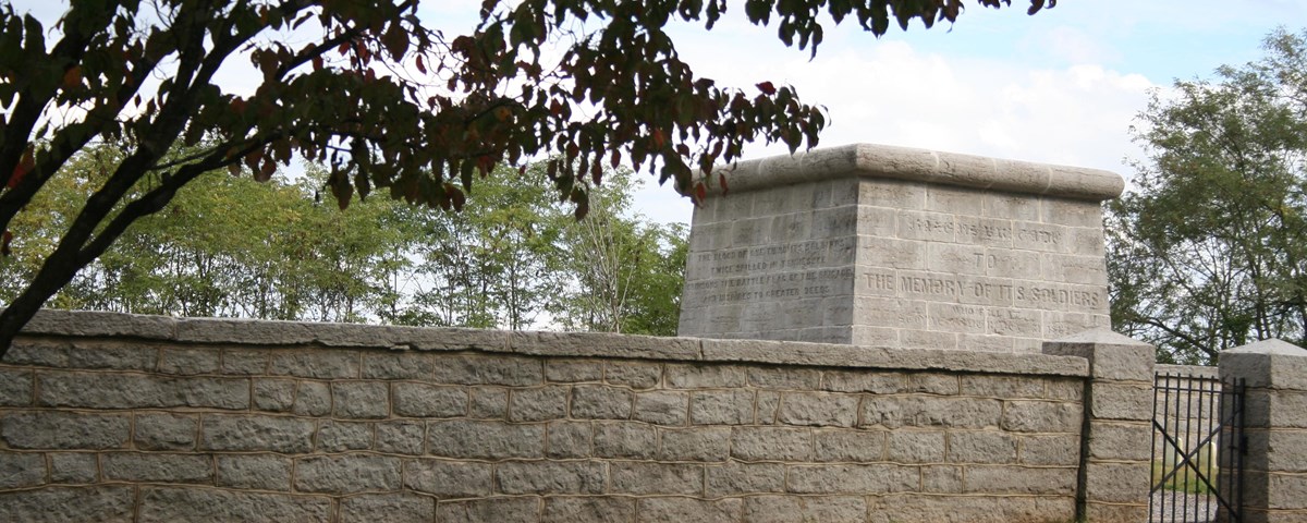 A grey stone wall and iron gate in front of a rectangular stone structure. A tree is in the foreground.