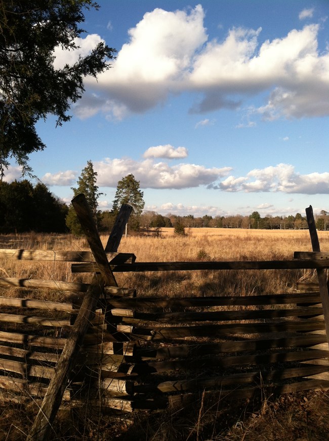 View of a field from behind a snake rail fence. Grass is brown and sky is blue with clouds.