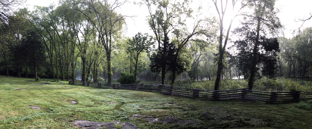 Fog shrouds a winding river at the base of a grassy slope. A snake rail fence runs through the mid ground dividing mowed grass to the front from taller grasses behind