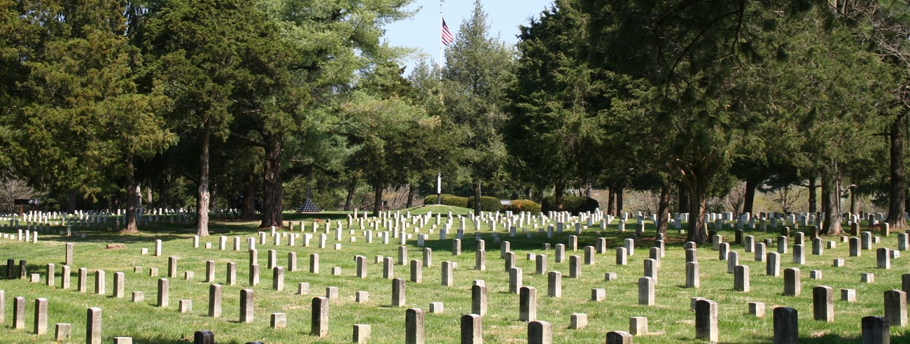 A white flagpole with an American Flag flies over a cemetery with rows of white headstones.