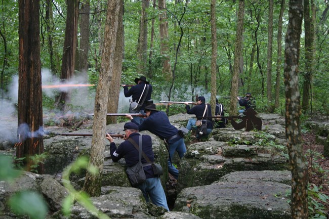 Living historians portraying Union soldiers fire muskets from behind large rock outcroppings.