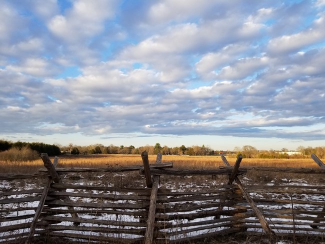 A field of brown grass dotted with patches of snow stretches behind a rail fence.