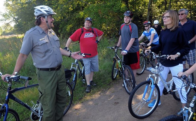 A park ranger wearing a bike helmet leans on a bicycle and speaks to a group of bicyclists.