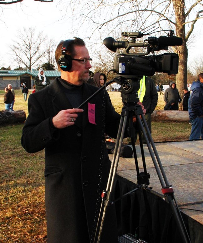A man in a long black overcoat stand behind a video camera on a tripod.