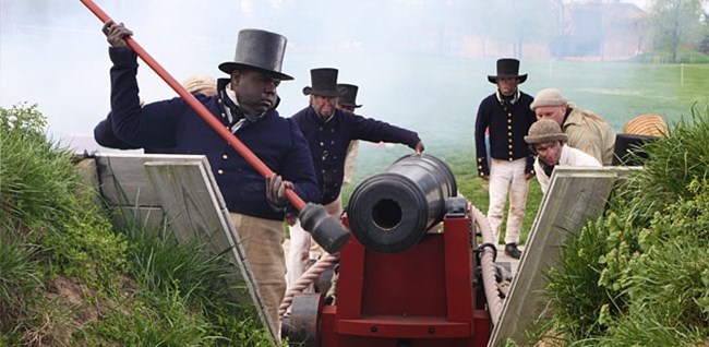 Photograph featuring living historians in period clothing loading a cannon.