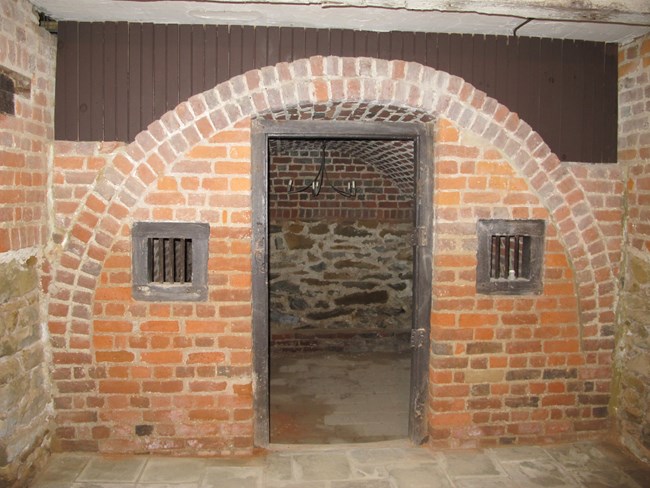 Photograph of a interior brick wall with a doorway in the middle and two small windows on either side.