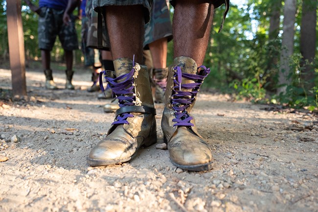 a dark-skinned man's feet in gold boots with purple laces along a dusty path