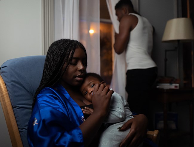 an African American mother holds her young child as the father looks out the curtained window