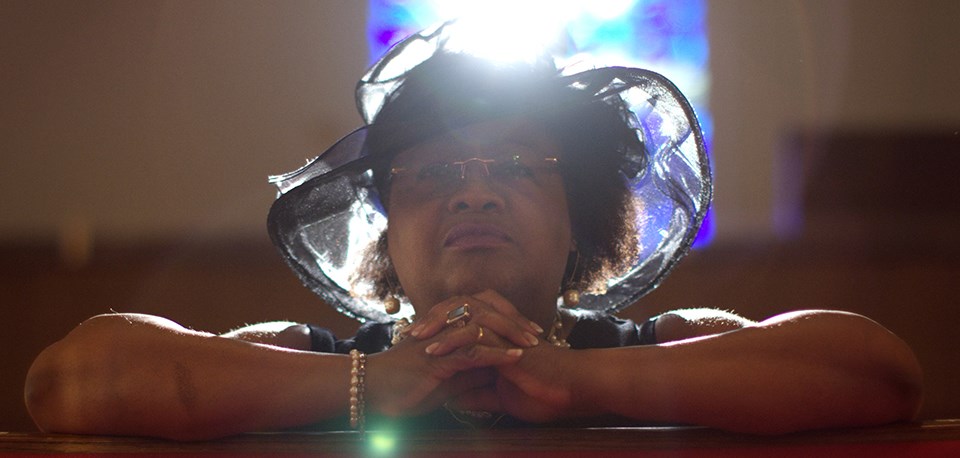 a woman in a hat in a church pew with sunlight streaming in the stained glass window behind her