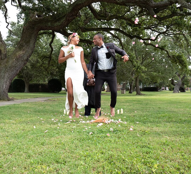 a young African American bride and groom jump over a broom under a large, old tree