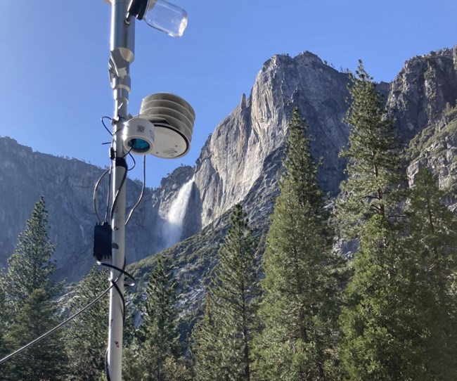 PurpleAir sensor attached to pole next to other monitoring devices with a background of a rock formation and waterfall.