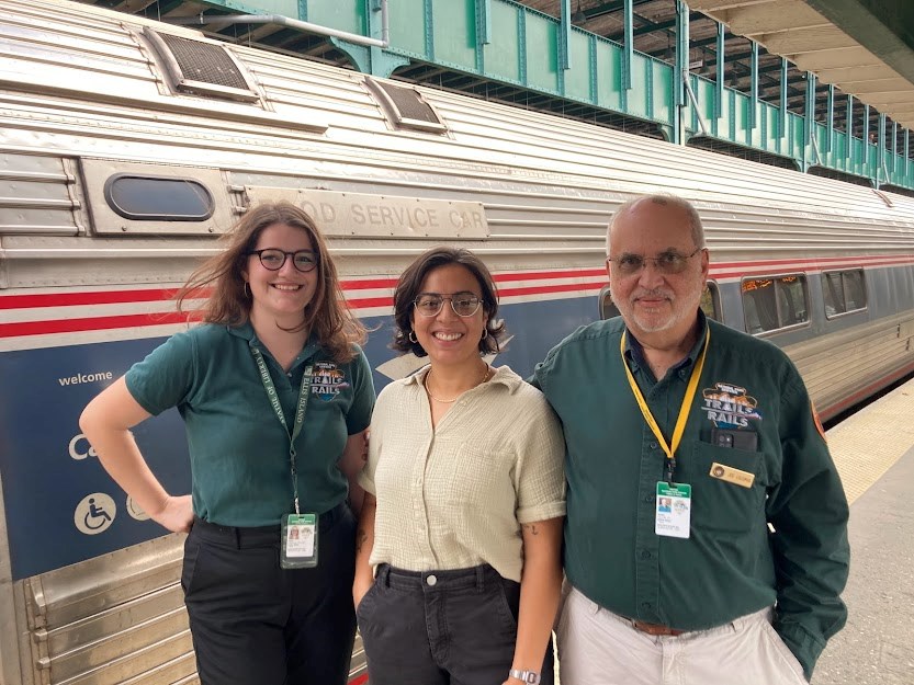 Three people stand in a line at an outdoor train station smiling for the camera. Behind them is a steel Amtrak train