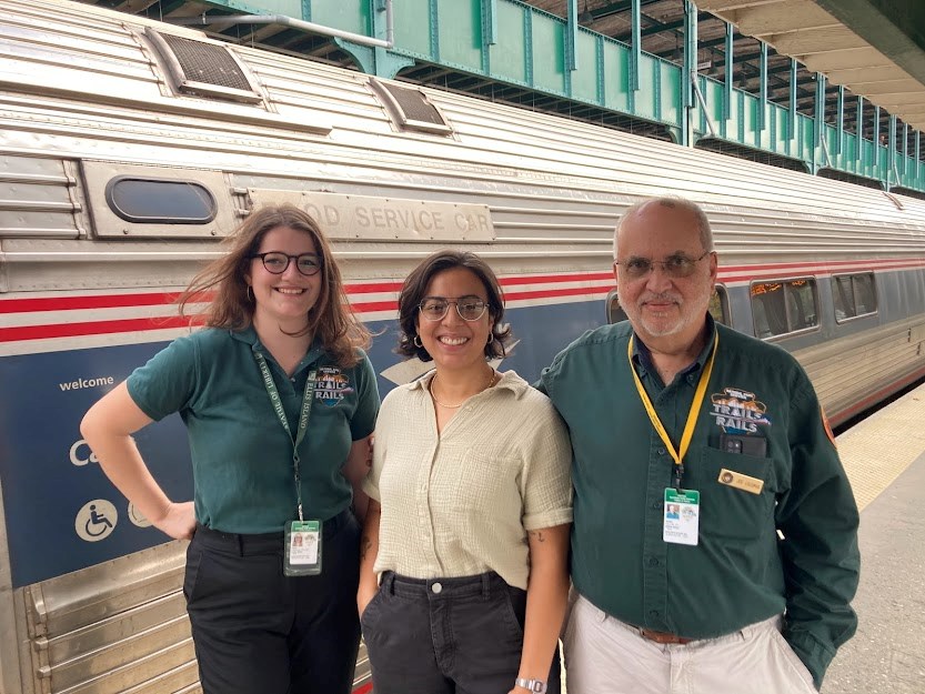 Volunteers on the New York Trails&Rails route. Three people stand in a line at an outdoor train station smiling for the camera. Behind them is a steel Amtrak train