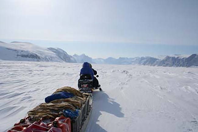 Two people on a snowmachine travel across the ice with a trailer of provision.