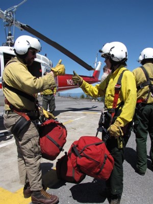 Two people stand facing each other, holding thumbs up, wearing white helmets and yellow shirts