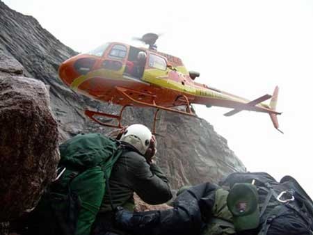 A helicopter hovers above several people next to a rocky cliff