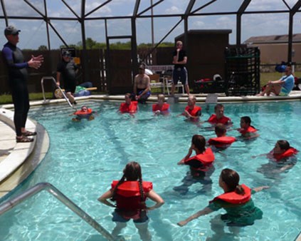 Water Ditching training Several people in orange life jackets float in a pool while looking up at a man standing on the edge of pool
