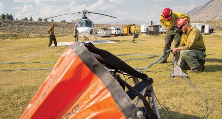 Two helicopter crew members hook up a water bucket to a cable with a chopper in the back