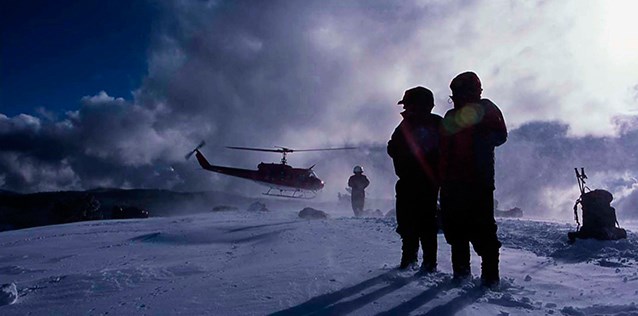 Two people stand in the foreground while a helicopter lands in the background