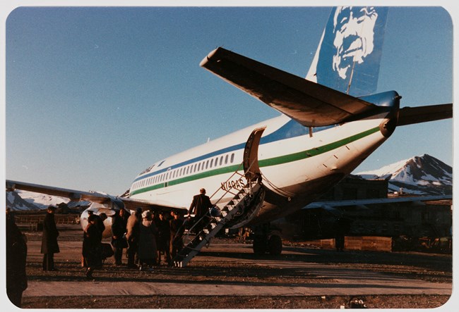 A group of people are boarding an airplane through the rear staircase. The airplane is an Alaska Airlines plane with the face logo painted on the tail.