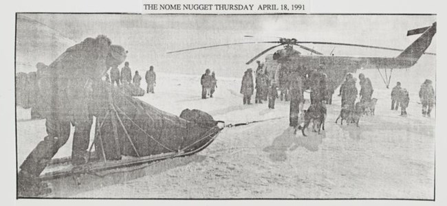 Wales Helicopters A black and white newspaper clipping from the Nome Nugget, showing dogs and a musher around approach a large helicopter.