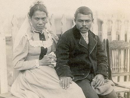 bride and bridegroom sit on a bench with a village street behind them