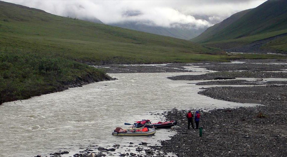 two people with inflatable kayaks on the bank of a river with mountains in the background