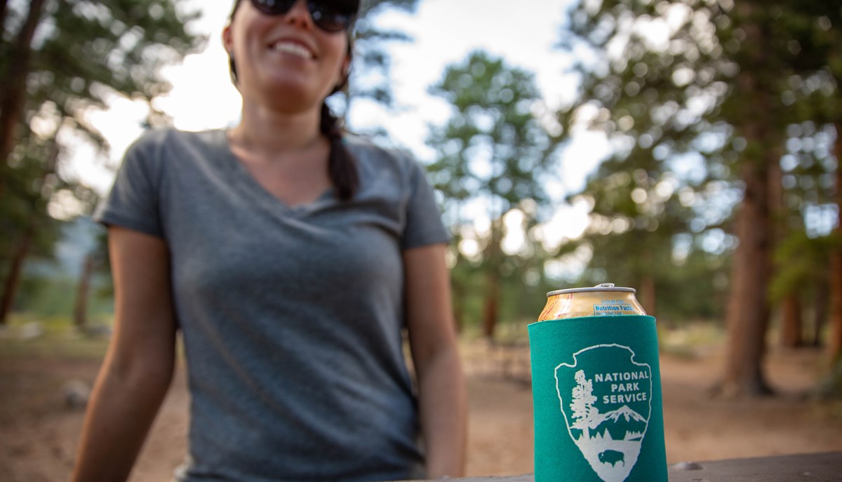 young woman sitting at a picnic table outside with the park service arrowhead on display on a coozie covering a can of sparkling water