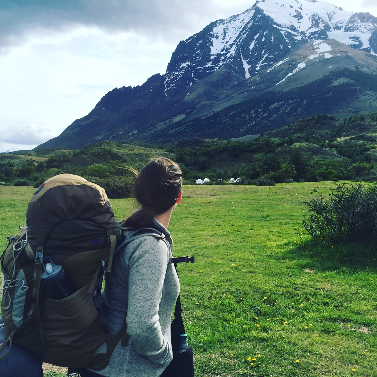 a woman wearing a large backpacking backpack looks up a tall snowy mountain