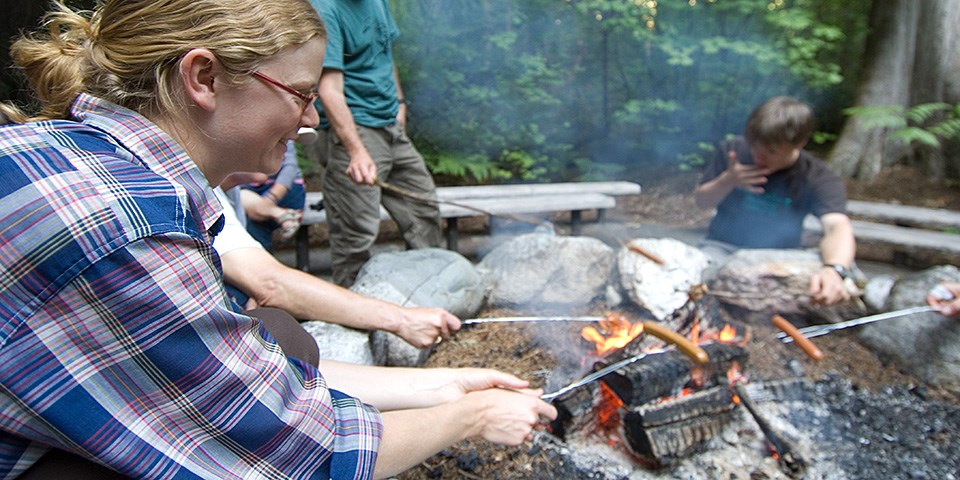 Youth site around a campfire cooking hot dogs