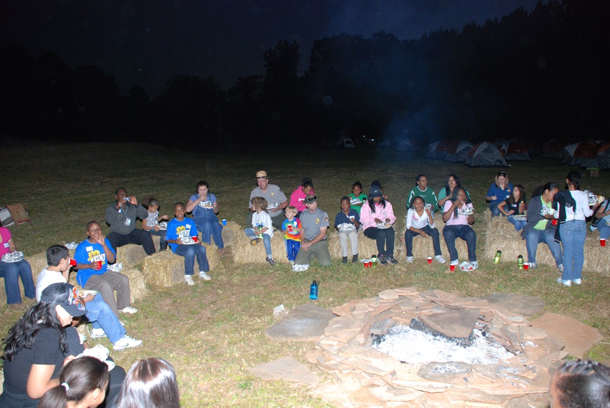 a group of kids, parents, and rangers sit around a large fire outside