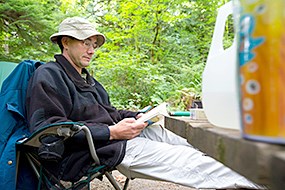 A man sits in a camp chair by a picnic table reading a book
