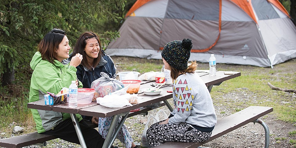 Three women sit at a picnic table eating and laughing. A tent is in the background.