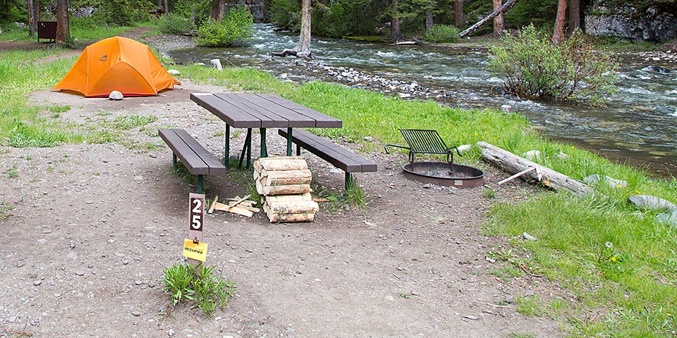 A campsite with a site marker, picnic table, fire ring, firewood and an orange tent