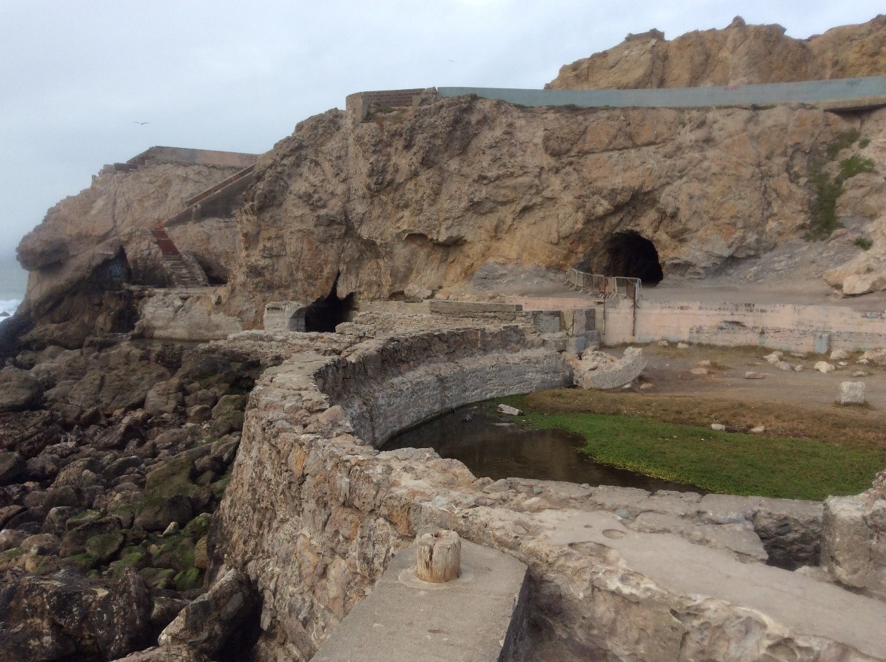 photo of a rocky seashore and cliffs with the remains of a walled swimming pool and 2 tunnels