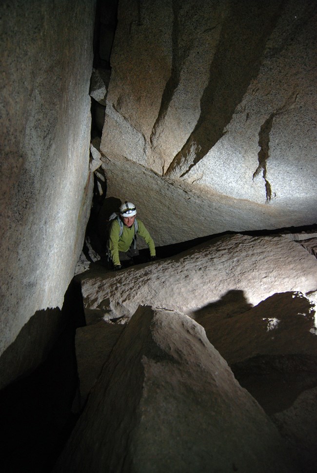 A photograph of a person wearing a helmet is on hands and knees exploring a room within a talus cave which is formed between large car to bus sized angular blocks of stone.