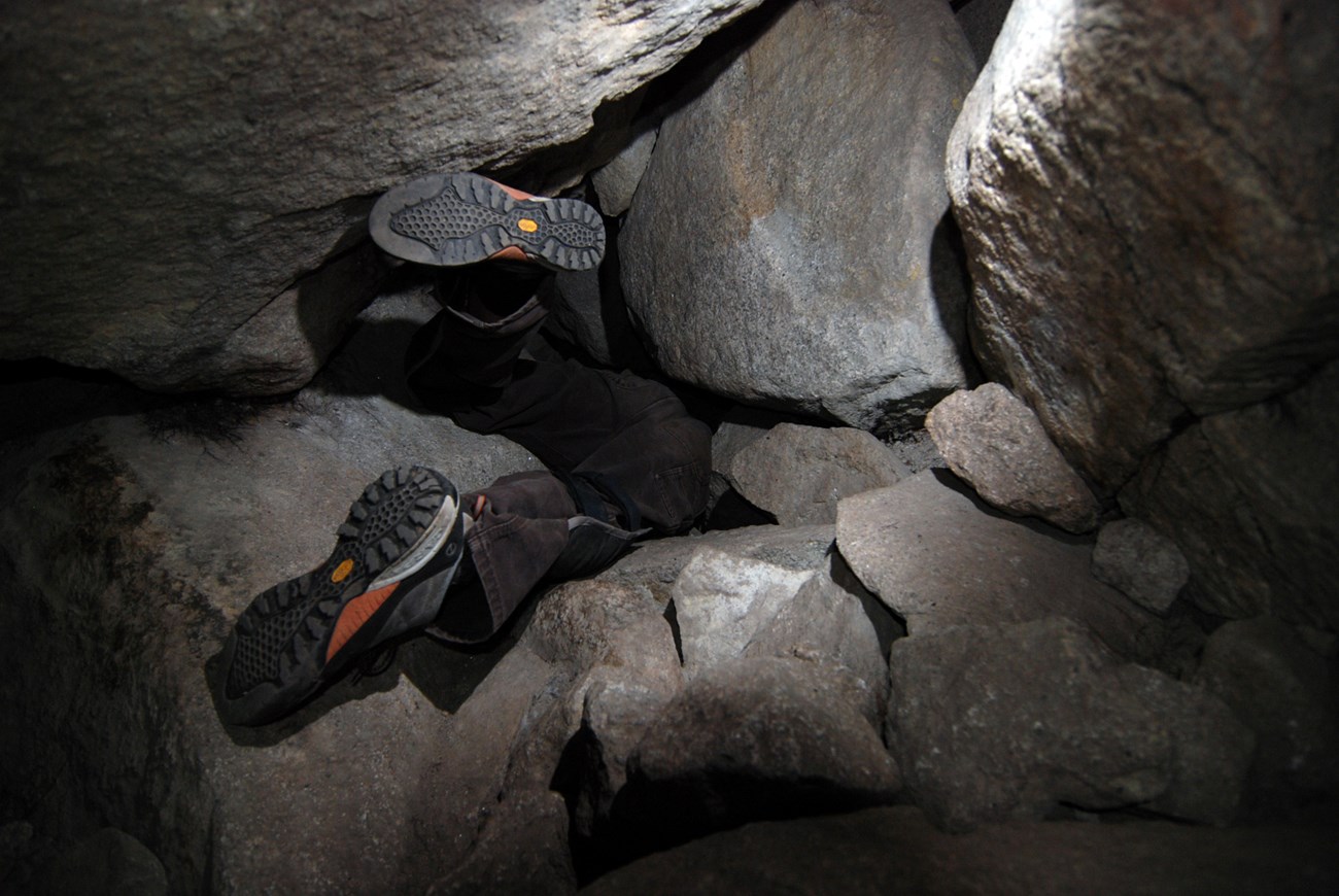 Photograph of a person’s shoes as they attempt to crawl through a tight passage between large boulders within a talus cave.