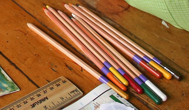 Colored pencils on a wood table next to a ruler and the edge of a green shirt sleeve
