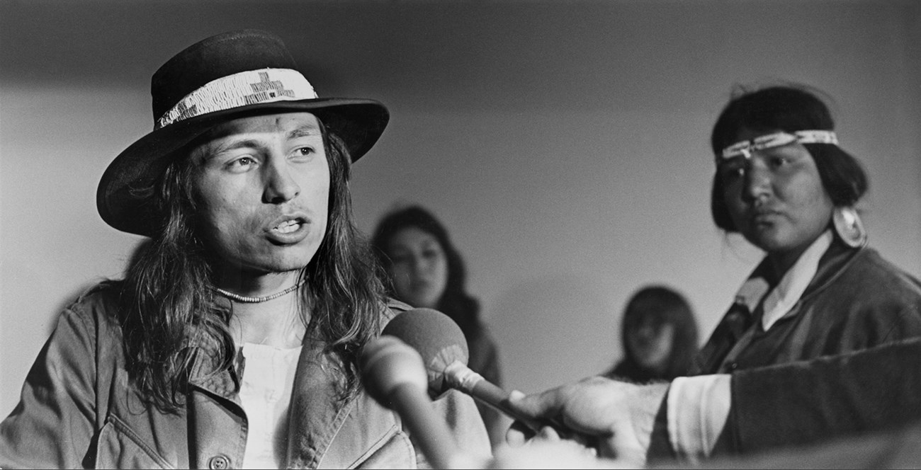 John Trudell and other occupiers at the press conference in the Senator Hotel on Ellis St. in San Francisco. A young male wearing broad-brimmed hat with beaded band stands before two microphones