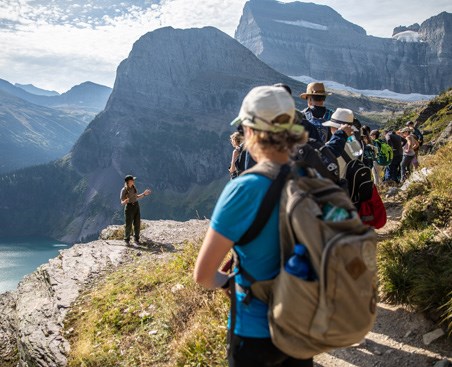 lt= Visitors on trail listen to ranger standing on cliff edge above a mountain valley with lake