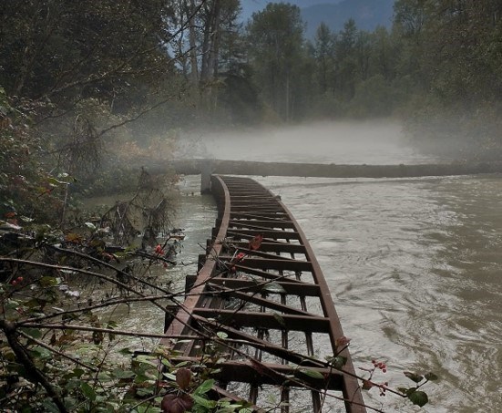 A damaged wooden bridge on a stormy day, partially crumbling into a forest-lined river.