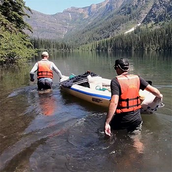 Two men wade in a forested mountain lake while pushing an inflatable gray boat
